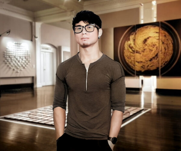 Portrait_1 Shohei Katayama standing in a gallery, wearing glasses and a dark henley shirt, with contemporary art pieces displayed on the walls behind him.