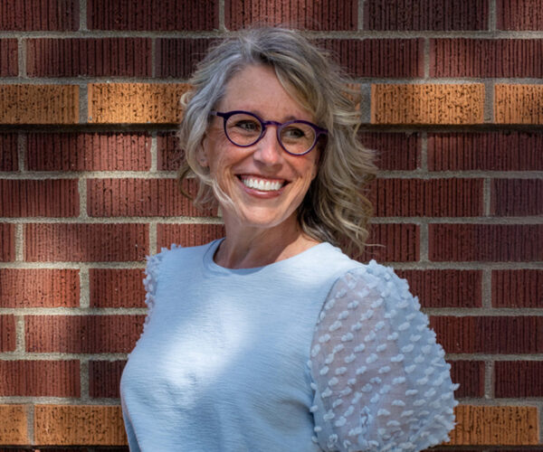 Richter-headshot Portrait of Jennifer Richter smiling warmly, wearing purple-rimmed glasses and a light blue blouse with textured sleeves, standing against a red brick wall in dappled sunlight.