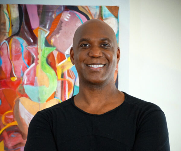 Wendell2228 Portrait of Wendell Smith standing in front of one of his colorful abstract paintings. He is smiling, with arms crossed, wearing a black shirt. The artwork behind him features layered organic shapes and bright colors, complementing his vibrant artistic style.