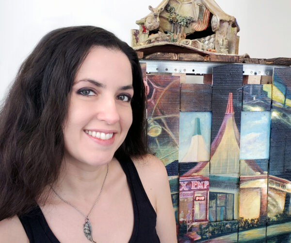 aclyman Portrait of Angelica Clyman, smiling with long dark hair, wearing a black tank top and a wing-shaped pendant necklace, standing in front of her artwork featuring architectural and landscape imagery painted on wood panels.