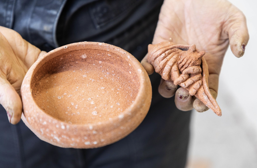 Close-up of a person's hands holding two small terracotta-colored sculptures made from algae-based clay—one a smooth round bowl and the other an organic, spiral-shaped form—highlighting the earthy texture and artistic potential of sustainable ceramics.