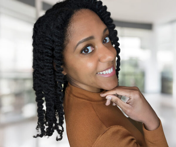 cephra A smiling woman with natural curls, wearing a rust-colored turtleneck and looking back over her shoulder. She poses with her hand near her chin against a softly blurred indoor background.