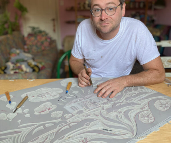 charlie spitzack Artist Charlie Spitzack in his studio, wearing glasses and a white T-shirt, seated at a table carving into a large linoleum block for printmaking.