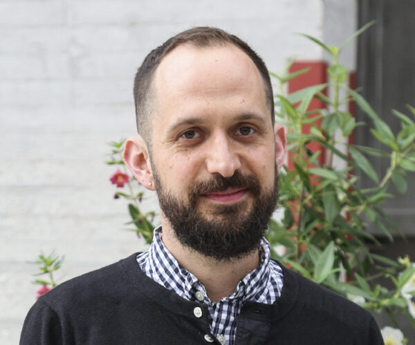 gilbert-headshot Portrait of artist James Gilbert wearing a checkered shirt and black sweater, standing outdoors in front of plants and a light-colored wall.