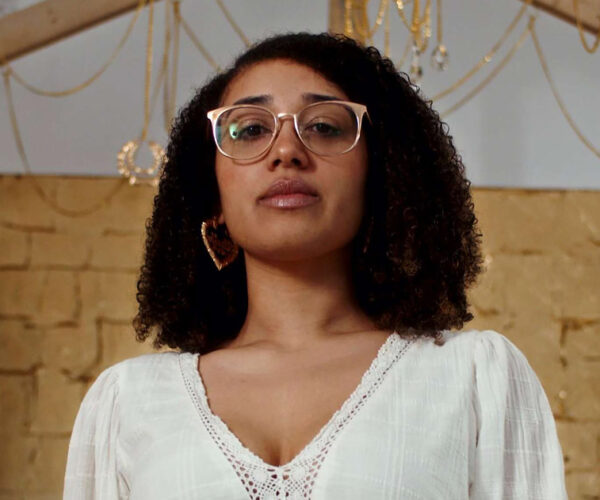 headshot_sydney_300-dpi Portrait of artist Sydney Maubert standing inside her installation Queen of the Swamp / PoeLilRichGirl, wearing a white blouse and patterned skirt, framed by gold-leaf walls and wooden beams.