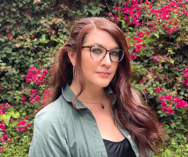 hopkins-headshot Portrait of artist Natali Hopkins against a nature background, wearing glasses and a dark top, looking directly at the camera with a neutral expression.