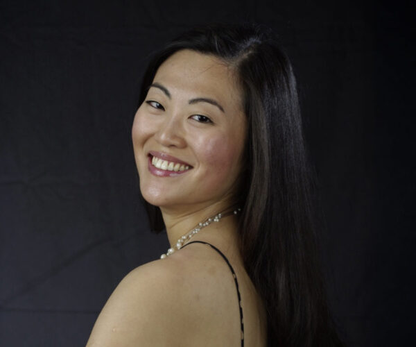jiay Portrait of pianist Jiayen Shen smiling over her shoulder, wearing a pearl necklace and black dress against a dark background