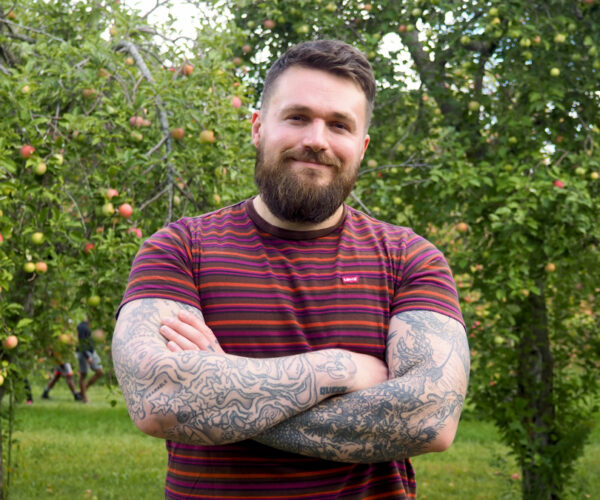 stedman Chris Stedman standing outdoors in an orchard, smiling with arms crossed. He wears a striped t-shirt and has tattooed arms, a beard, and short hair, with apple trees filling the background.