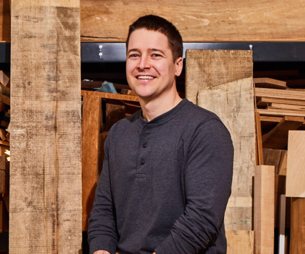 tinkham Portrait of artist Spencer Tinkham seated in a woodshop, smiling at the camera. He wears a dark long-sleeve shirt and is surrounded by stacks of cut and raw wood in the background.