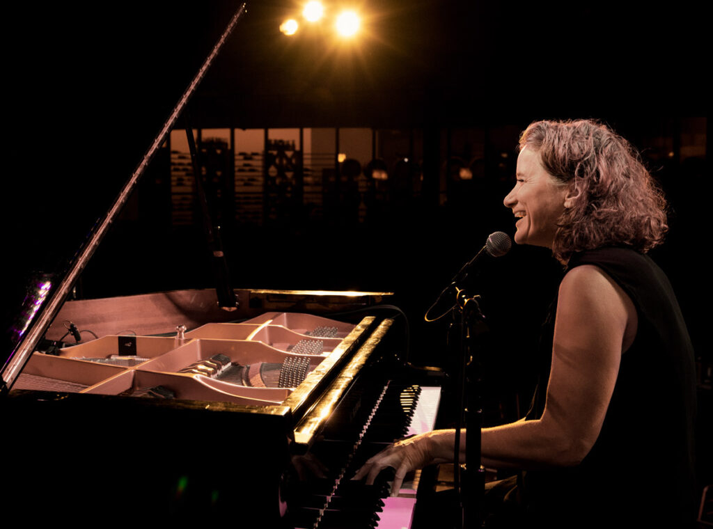 Musician Susan Werner performs on stage, smiling as she sings into a microphone while playing a grand piano under warm stage lights.