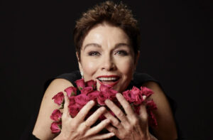 Portrait of cabaret performer Christine Andreas smiling brightly and holding a bouquet of red roses close to her face against a black background.