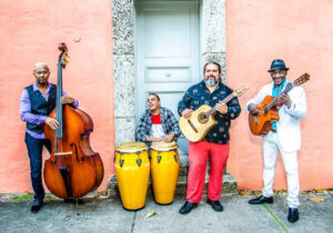 A four-piece Latin band stands against a peach-colored wall, featuring a double bassist, percussionist with congas, and two guitarists, all smiling and ready to perform.