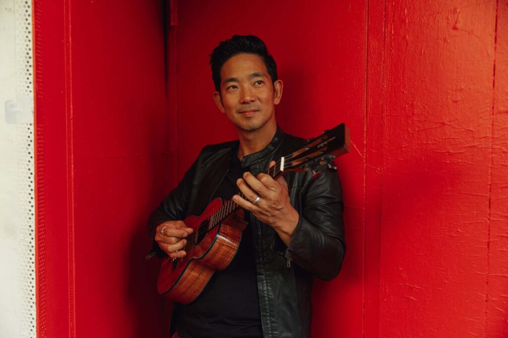 Ukulele virtuoso Jake Shimabukuro stands against a vivid red wall, strumming his instrument with a relaxed smile. Dressed in a black leather jacket and dark shirt, he radiates calm confidence and creative energy, embodying the boundary-breaking spirit that has made him one of the world’s most celebrated ukulele players.