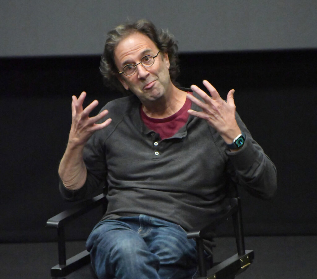 Writer and speaker Danny Rubin gestures expressively with both hands while sitting on a director’s chair, wearing glasses and a dark pullover over a red shirt.