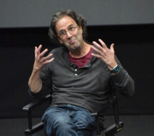 Writer and speaker Danny Rubin gestures expressively with both hands while sitting on a director’s chair, wearing glasses and a dark pullover over a red shirt.