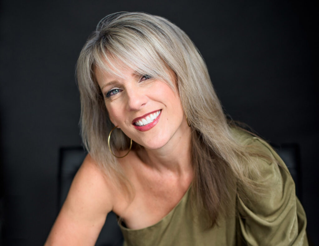 Portrait of Darcy Cooke smiling warmly against a dark background. She wears a one-shoulder olive green top and large gold hoop earrings, her blonde hair softly framing her face.