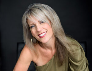 Portrait of Darcy Cooke smiling warmly against a dark background. She wears a one-shoulder olive green top and large gold hoop earrings, her blonde hair softly framing her face.