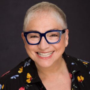 Portrait of Debra Barsha smiling warmly against a dark background. She wears bold blue glasses and a black shirt patterned with colorful flowers, exuding creativity and confidence.
