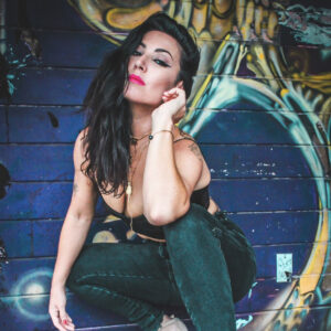 A woman in a confident pose against a colorful graffiti wall, wearing bold makeup and a black outfit, with bright pink lipstick and a direct gaze.