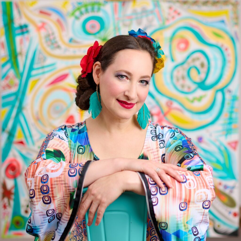 Singer-songwriter Rachael Sage poses against a colorful, abstract backdrop. She wears bright, patterned clothing, turquoise earrings, and flowers in her hair, radiating creativity and joyful energy.