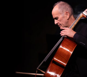 Cellist Carter Brey performs on stage, focused intently on playing his cello. The background is dark, highlighting the warm wood tones of the instrument.