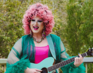 A drag performer with voluminous pink curls and bold makeup plays a turquoise acoustic guitar outdoors, wearing a green ruffled robe over a hot pink outfit and smiling brightly.