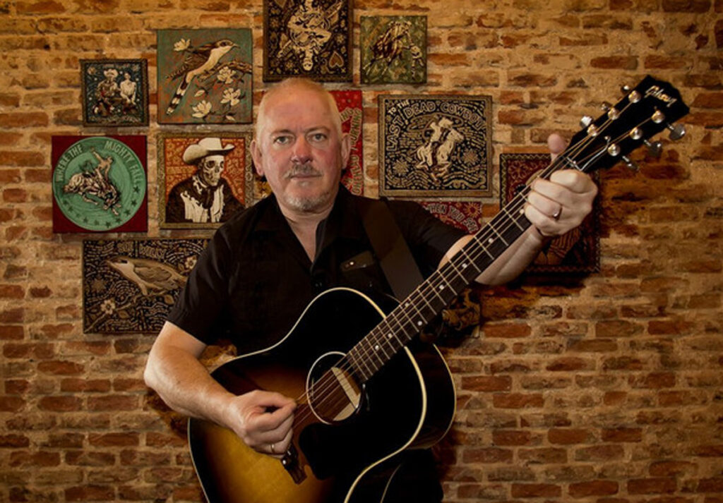 Musician and artist Jon Langford stands before a brick wall decorated with his own artwork, holding an acoustic guitar mid-strum. His serious expression contrasts with the vivid, folk-inspired imagery behind him, capturing the creative fusion of punk energy and Americana roots that defines his work.