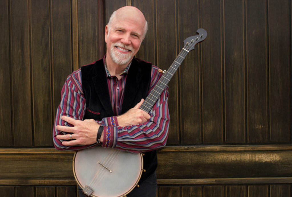 Folk musician John McCutcheon smiles warmly while holding a banjo across his chest. He wears a striped shirt and dark vest, standing before a wooden backdrop that evokes the roots and warmth of traditional folk music.