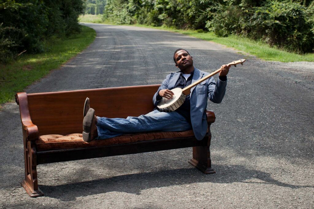 A man lounges on a wooden church pew placed in the middle of a rural road, playing a banjo. He gazes thoughtfully into the distance, blending a sense of history, music, and storytelling in a quiet, reflective setting surrounded by greenery.