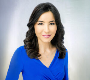 Journalist Roxana Saberi poses for a professional headshot wearing a royal blue top and a delicate necklace, smiling softly against a light gradient background.