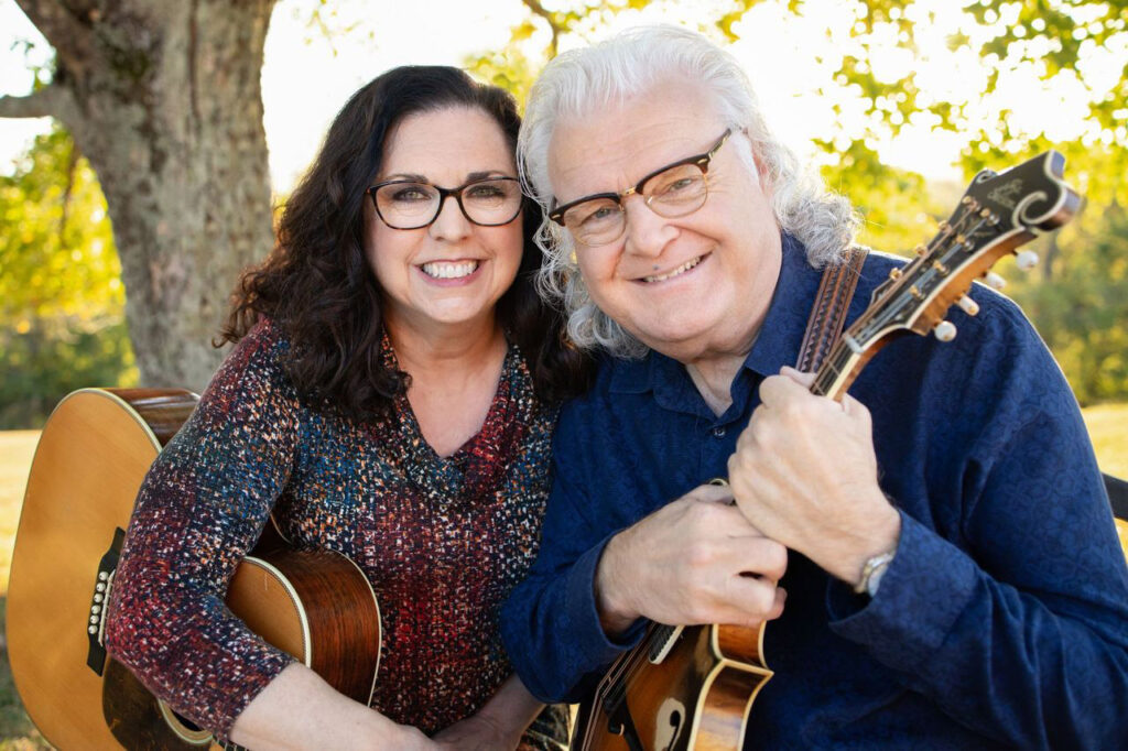 Ricky Skaggs and Sharon White smile warmly outdoors, each holding their instruments — Skaggs with his mandolin, White with her guitar. Sunlight filters through the trees behind them, capturing the warmth, harmony, and enduring partnership that define their decades-long musical and personal journey.