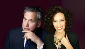 Side-by-side portrait of Billy Stritch in a navy suit and Gabrielle Stravelli wearing a statement necklace and smiling warmly against a dark plum background.