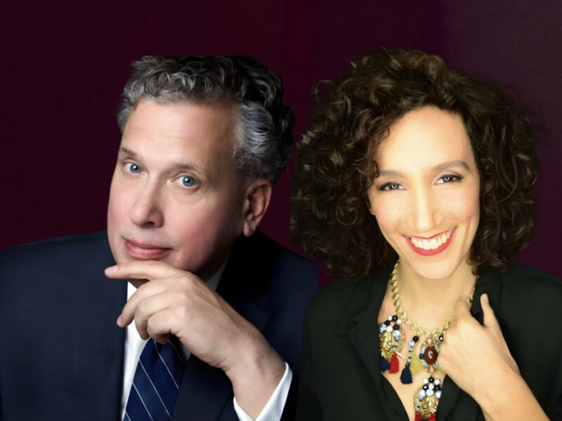 Side-by-side portrait of Billy Stritch in a navy suit and Gabrielle Stravelli wearing a statement necklace and smiling warmly against a dark plum background.