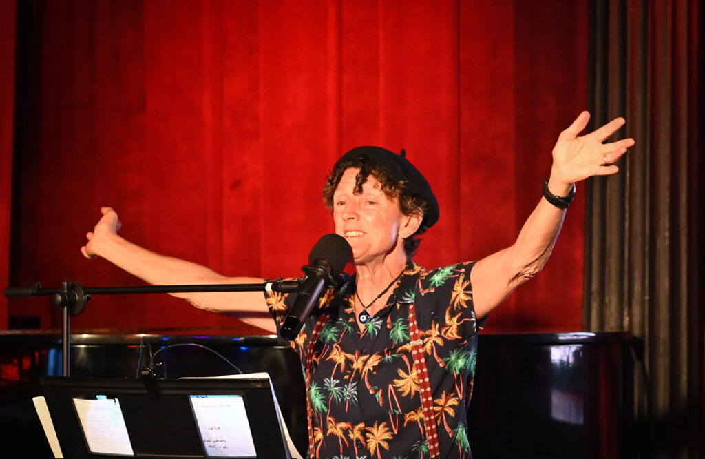 A performer wearing a tropical-print shirt and black beret sings and plays the piano on stage with arms outstretched in front of a red curtain.