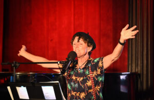 A performer wearing a tropical-print shirt and black beret sings and plays the piano on stage with arms outstretched in front of a red curtain.