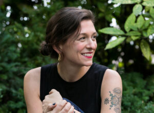 A woman with dark brown hair pulled into a loose bun sits outdoors against lush green foliage. She smiles brightly and looks to her left. She wears a black sleeveless top, gold hoop earrings, and has a floral tattoo on her upper arm. The mood is friendly and natural.