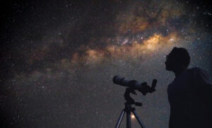 Silhouette of a person standing beside a telescope under a night sky filled with stars and the glowing band of the Milky Way stretching across the horizon.