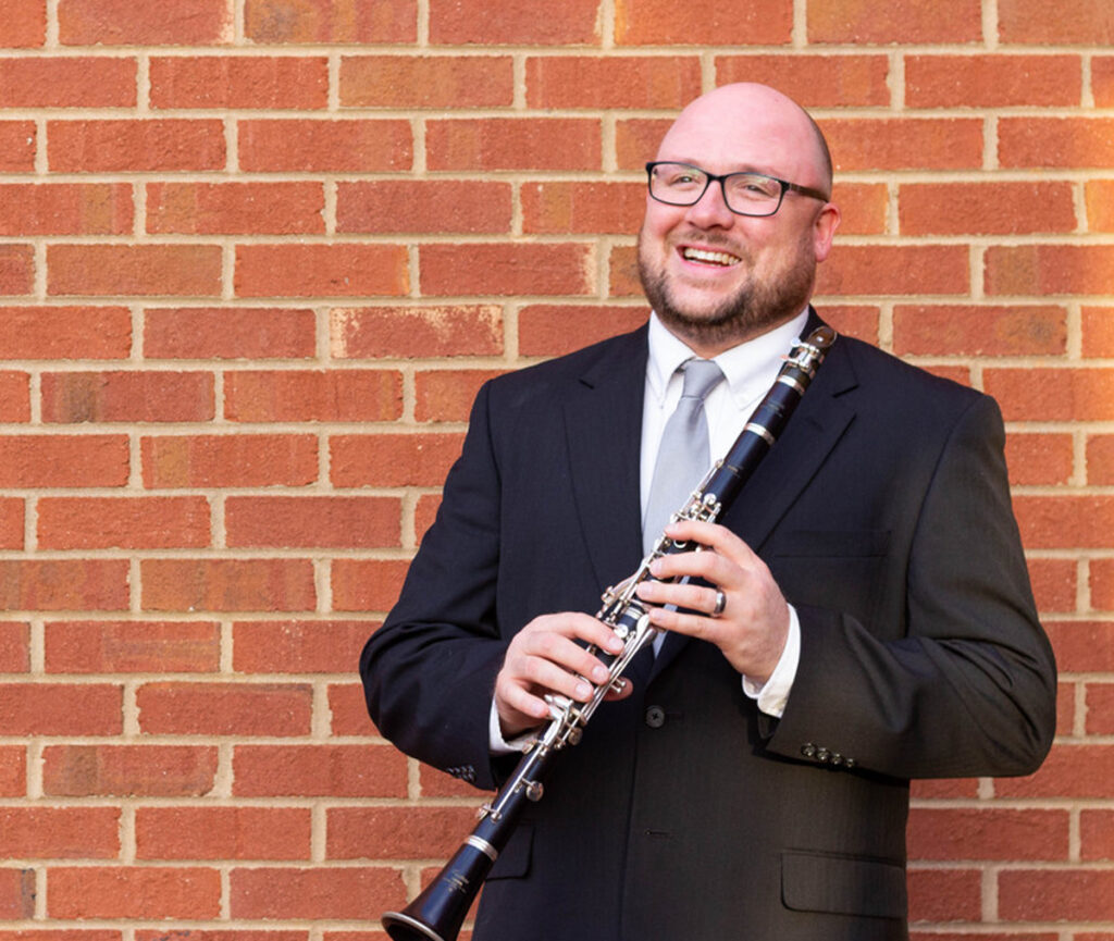A smiling man in a suit stands against a red brick wall, holding a clarinet. He wears glasses, a gray tie, and a wedding band. He looks slightly off camera, appearing friendly and approachable.