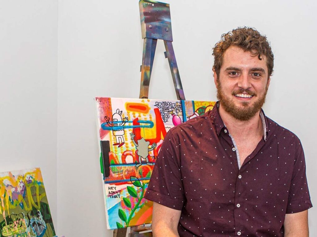 Man with curly hair and beard sitting beside bright abstract paintings on easels.