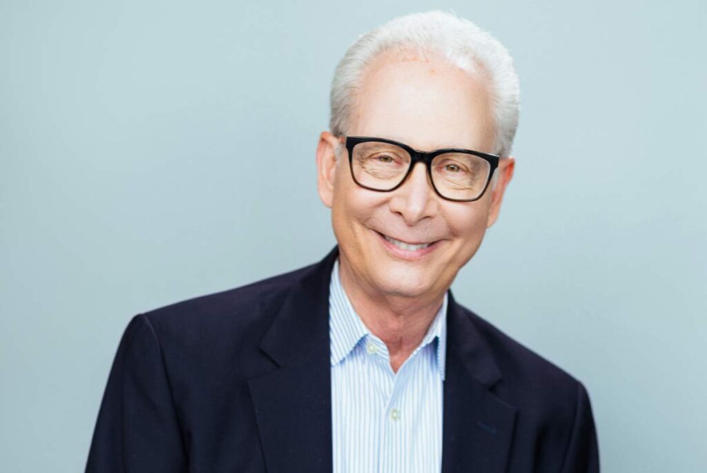Smiling man wearing glasses, a blue striped shirt, and navy blazer against a light background.