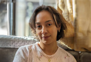 A young woman with short, softly curled brown hair sits indoors in warm natural light. She wears a pale pink sweater with small embroidered flowers, gold hoop earrings, and layered gold necklaces. She gazes calmly at the camera with a gentle, relaxed expression. The background is softly blurred.
