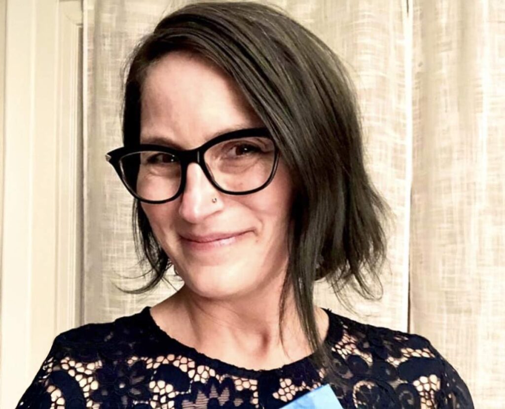 Smiling woman with short dark hair and glasses, wearing a navy lace top, standing in front of a softly lit curtain background.