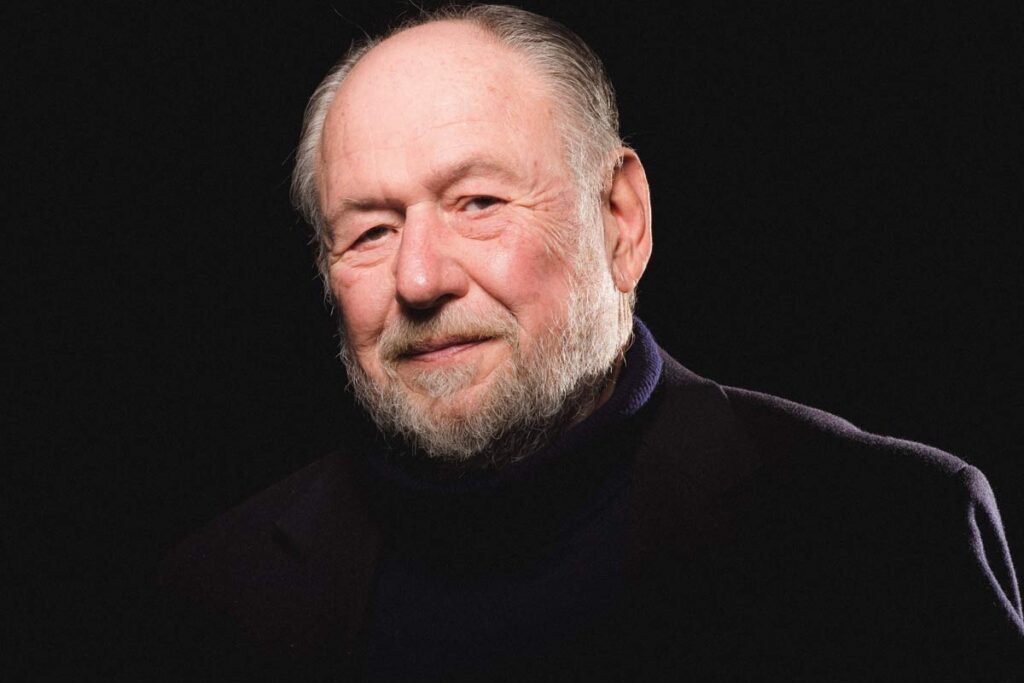 Headshot of an older man with a white beard and gentle smile, set against a black background.