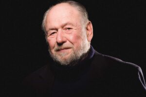 Headshot of an older man with a white beard and gentle smile, set against a black background.