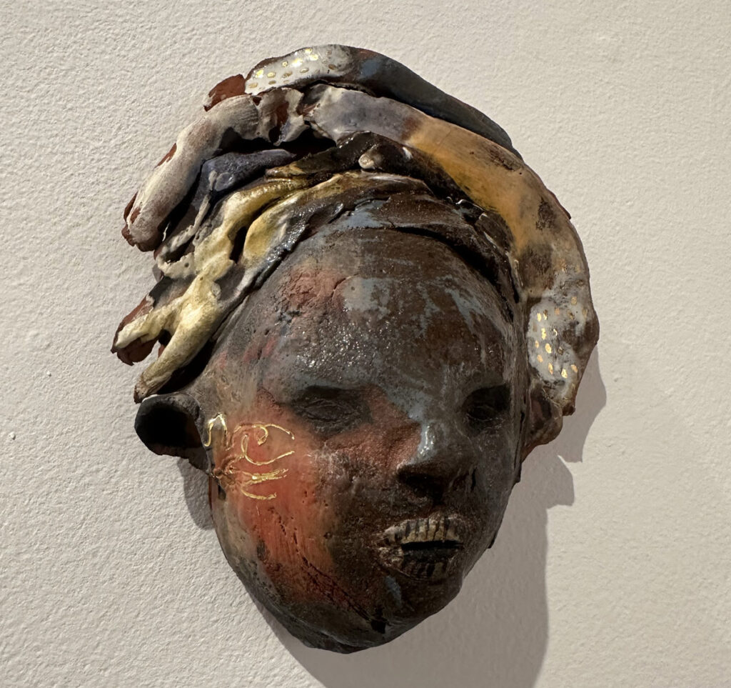 Textured ceramic wall sculpture of a face with layered earthy tones, detailed features, and organic shapes forming a headdress.