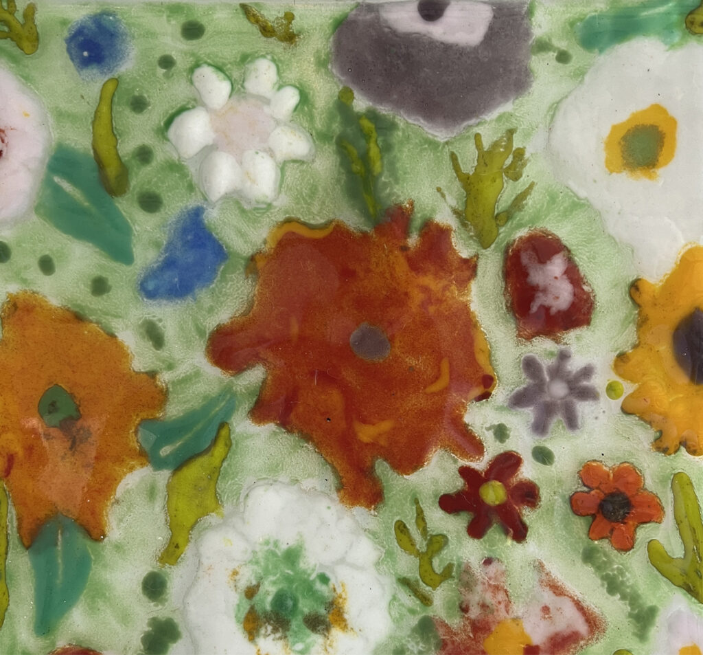 Close-up of fused glass artwork featuring colorful floral shapes in orange, red, white, and green on a translucent green background.