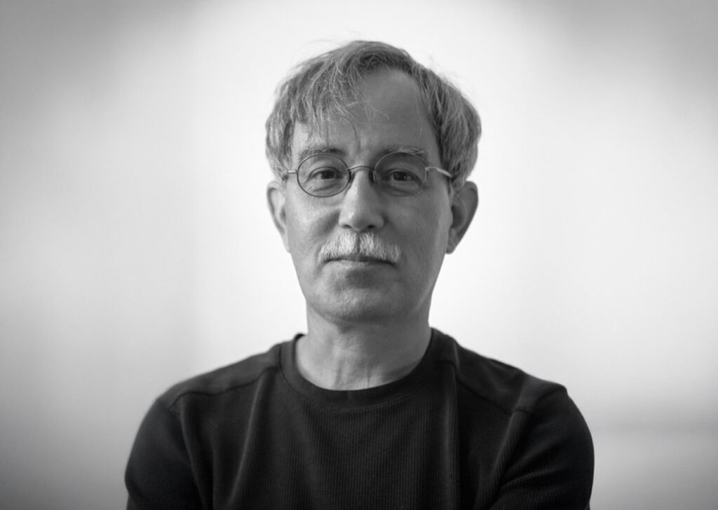 A grayscale portrait of an older man with glasses and a trimmed mustache. He wears a dark long-sleeve shirt and looks directly at the camera with a calm, introspective expression. The background is softly blurred.