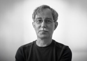 A grayscale portrait of an older man with glasses and a trimmed mustache. He wears a dark long-sleeve shirt and looks directly at the camera with a calm, introspective expression. The background is softly blurred.