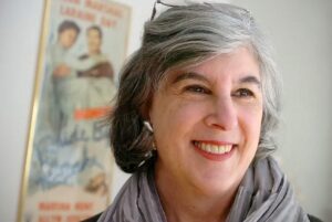 Smiling woman with gray hair and red lipstick, wearing a gray scarf with a vintage movie poster in the background.
