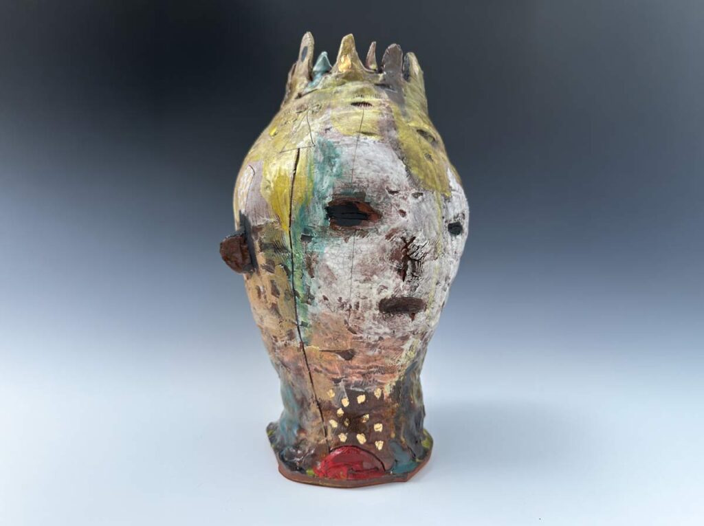 Textured ceramic sculpture of a head with muted colors of yellow, teal, and rust, featuring a crown-like top.
